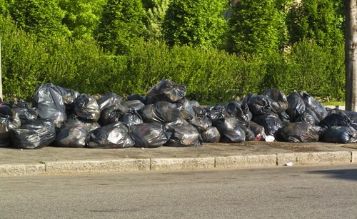 A long row of black plastic rubbish bags filled with waste material, stacked along the edge of a paved road. The bags are tightly packed and appear to contain household or garden refuse. Behind the bags, there is a dense line of green bushes and trees, indicating an outdoor setting possibly near a suburban or rural area in Tooting. The ground surface in front of the bags is a concrete or tarmac pavement, with a curb separating the waste from the roadway. The scene suggests an accumulation of rubbish awaiting collection or disposal, consistent with a professional waste removal service such as Commercial Waste Removal Tooting that handles domestic and commercial rubbish collection in the local area. Natural daylight highlights the textures and colors of the refuse bags, which are mostly matte, with some showing slight creases and folds due to handling or stacking.
