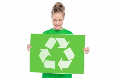 A young woman with blonde hair tied back in a ponytail, wearing a bright green long-sleeve shirt, is smiling and holding a large rectangular green sign featuring a white recycling symbol composed of three arrows forming a triangle. The background is plain white with no additional objects or scenery visible, emphasizing her cheerful expression and the environmentally themed message of recycling. This image is suitable for illustrating waste management or rubbish recycling initiatives, aligning with services like those offered by Commercial Waste Removal Tooting, a company operating within the SW17 postcode area. The focus on the recycling symbol highlights environmentally responsible waste disposal practices that are relevant to local rubbish removal services in Tooting.