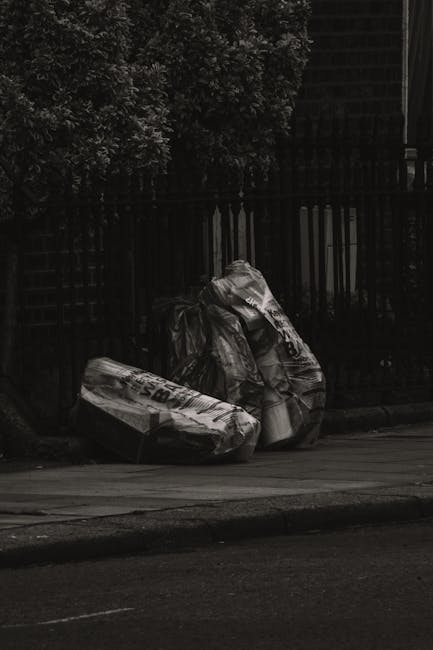 A woman with short dark hair, wearing a black T-shirt with white text and black pants, is bent over, actively disposing of waste into a tall, cylindrical stainless steel public trash bin situated on a